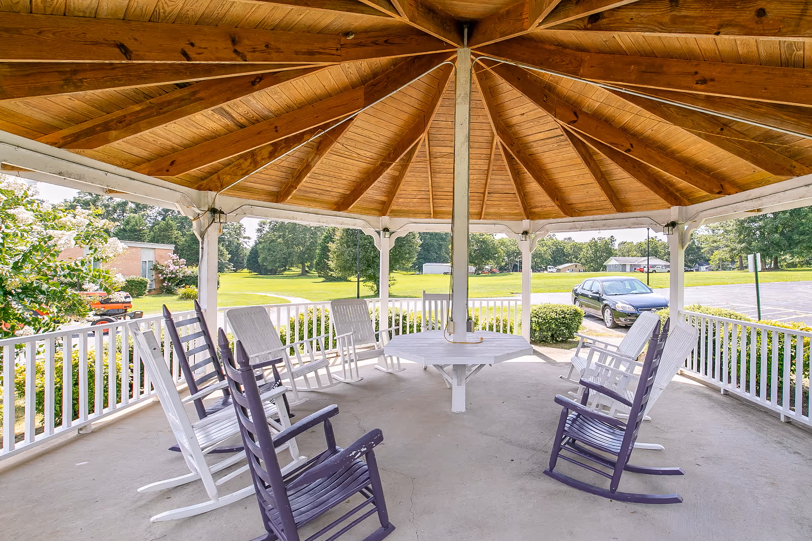 A wooden gazebo with a high peaked roof and white railing, furnished with white and purple rocking chairs arranged around a central table. The gazebo overlooks a green lawn, trees, a parking lot with a car, and nearby buildings under a clear sky.