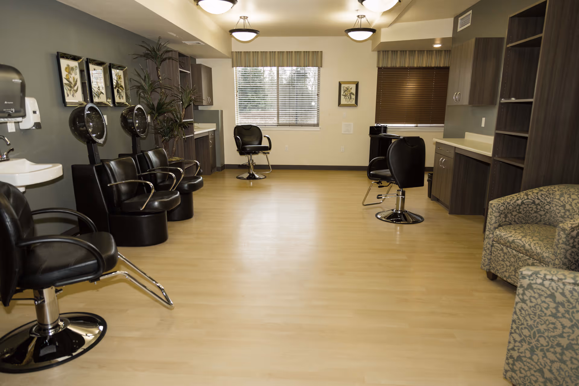 Interior view of a salon area in a senior living facility with several black salon chairs, hair dryers, cabinets, a sink, and two windows with blinds. There is also a patterned armchair on the right side.