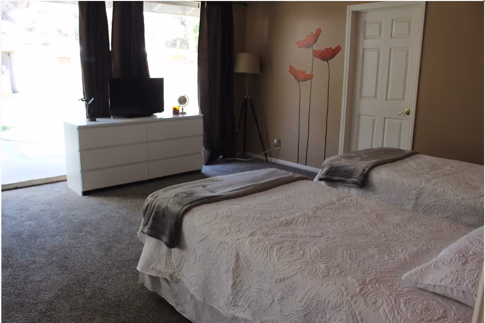 A bedroom with two twin beds covered in white patterned bedspreads and gray throws at the foot. A white dresser with a small TV and decorative items sits against the wall near a window with dark curtains. A floor lamp stands in the corner next to a wall decorated with red flower decals. A closed white door is visible on the right side.