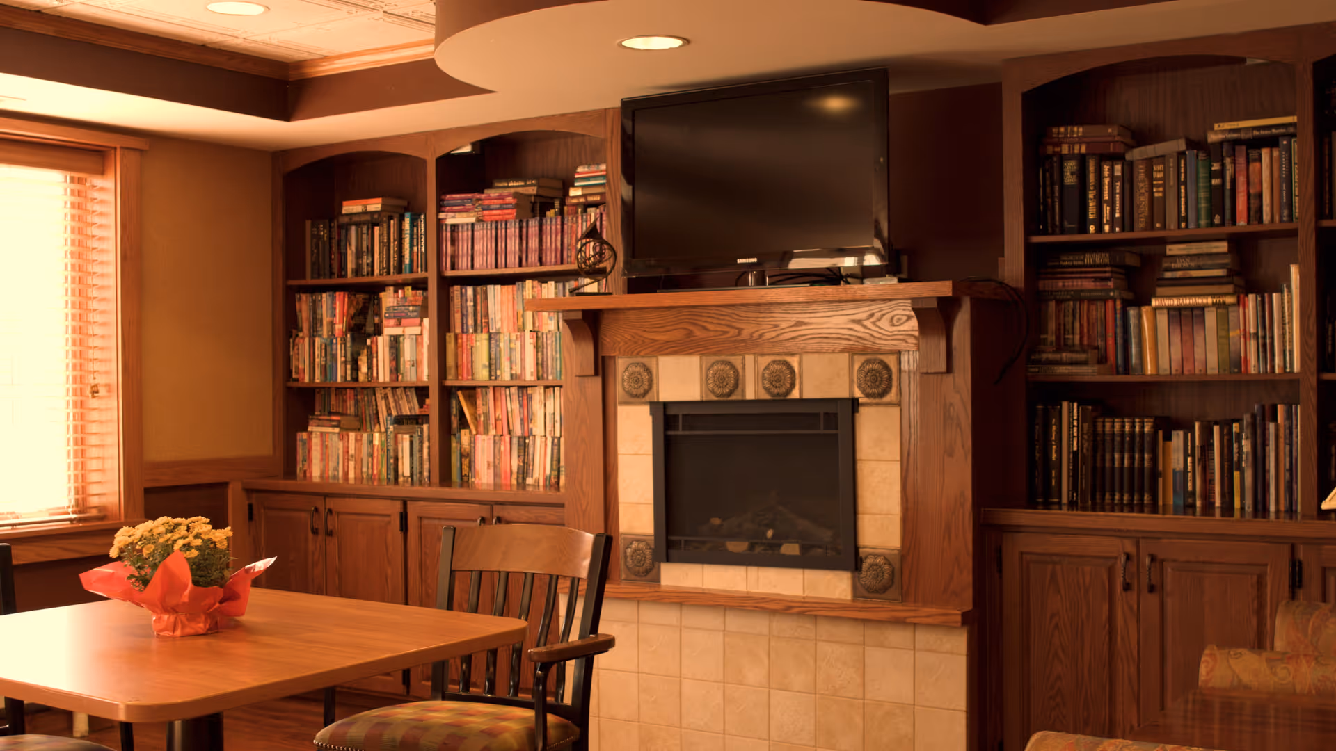 Cozy interior room with wooden bookshelves filled with books on either side of a fireplace topped with a flat-screen TV. A wooden table with a potted plant wrapped in red paper and a wooden chair are in the foreground. A window with blinds allows natural light into the room.