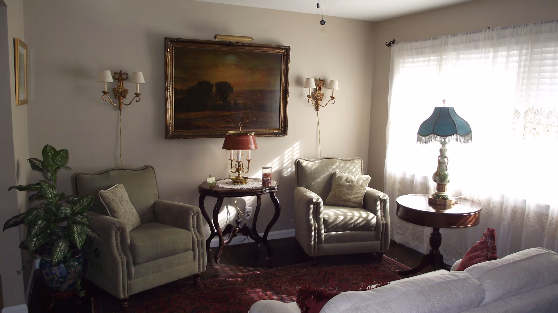 Sunlit living room with two upholstered armchairs, side tables with lamps, a framed painting, and a potted plant by a curtained window.