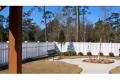 Enclosed outdoor courtyard with a white vinyl fence, concrete walkways, circular planting beds and a bench under a blue sky.