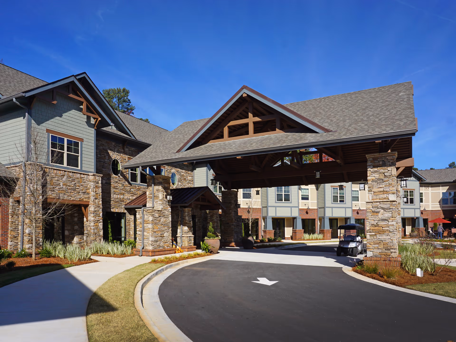 Exterior view of Village Park Peachtree Corners senior living facility showing a covered entrance with stone pillars, a driveway with an arrow painted on the asphalt, and a golf cart parked near the entrance. The building features stone and siding walls with multiple windows under a clear blue sky.