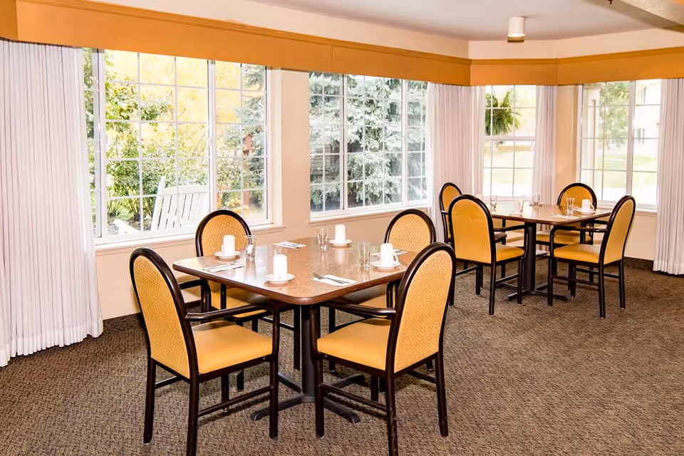 A bright dining room with two square tables, each surrounded by four yellow cushioned chairs. The tables are set with white cups, saucers, glasses, and silverware. Large windows with white curtains let in natural light and offer a view of green trees and a rocking chair outside.