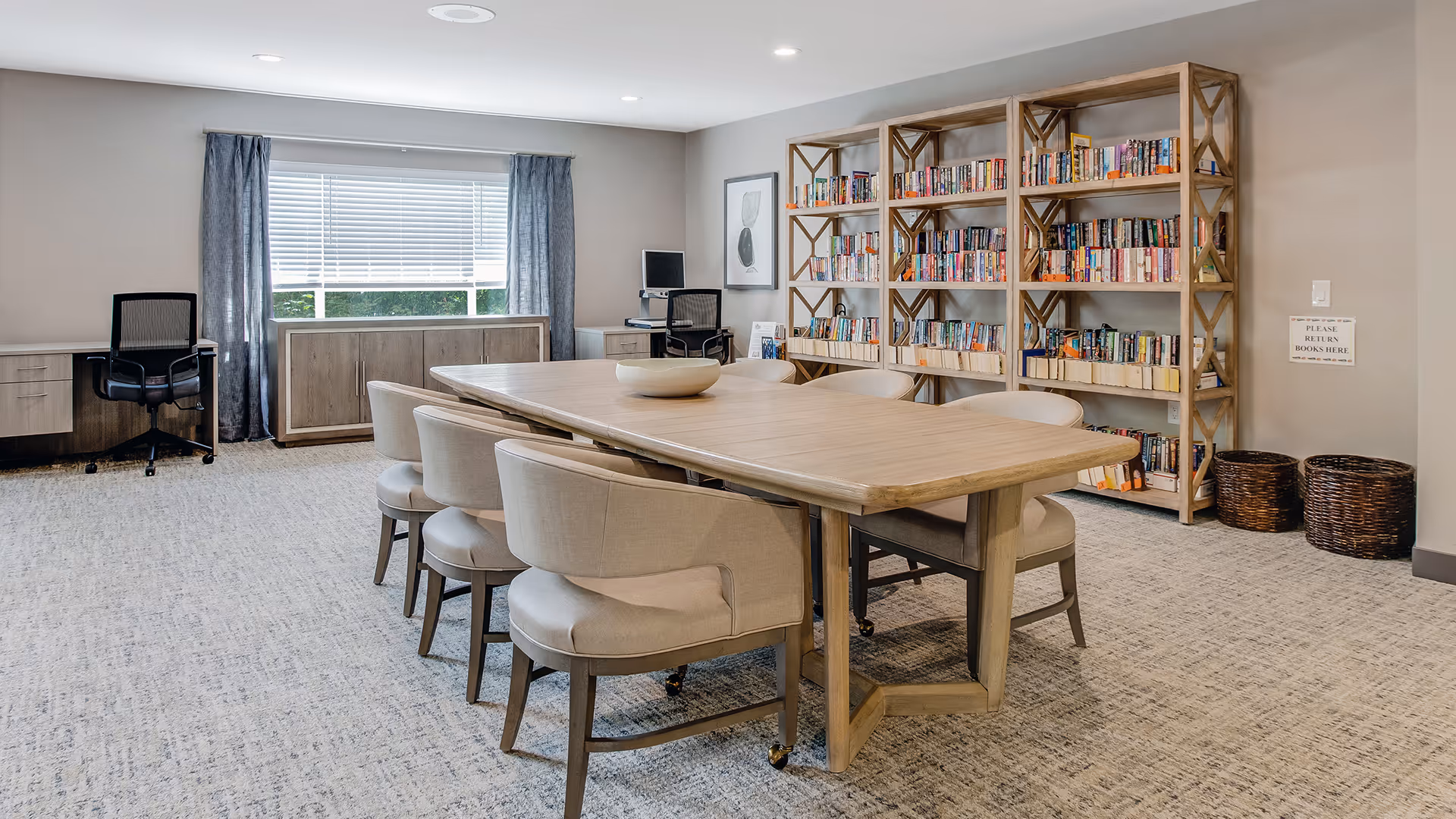 A well-lit room with a large wooden table surrounded by eight cushioned chairs. Against the far wall, there are multiple bookshelves filled with books. Two wicker baskets are placed near the bookshelves. A window with closed blinds and curtains is on the left wall, with two office chairs and desks beneath it. The room has a neutral color palette with beige walls and carpeted flooring.