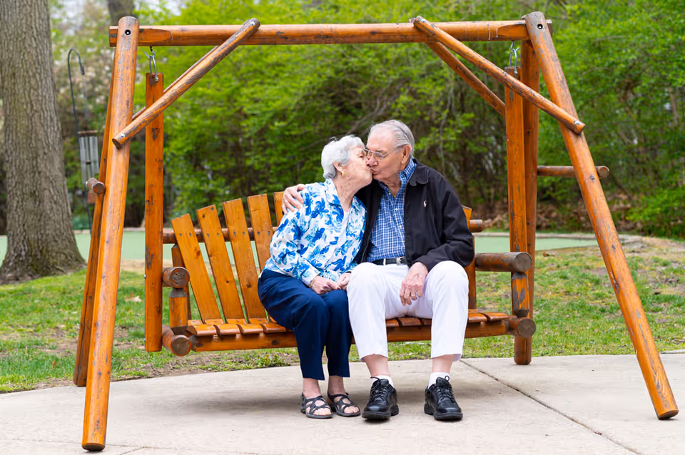 An elderly couple sitting on a wooden outdoor swing bench in a park-like setting. They are sharing a kiss, surrounded by green trees and grass.