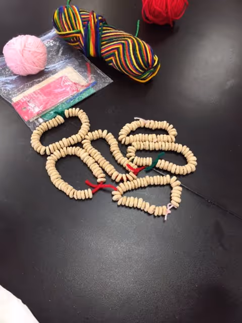 A black table surface with three balls of yarn in pink, multicolored, and red colors. There are several strings of cereal loops threaded onto yarn pieces in different colors laid out on the table. A small plastic bag with some items inside is also visible.