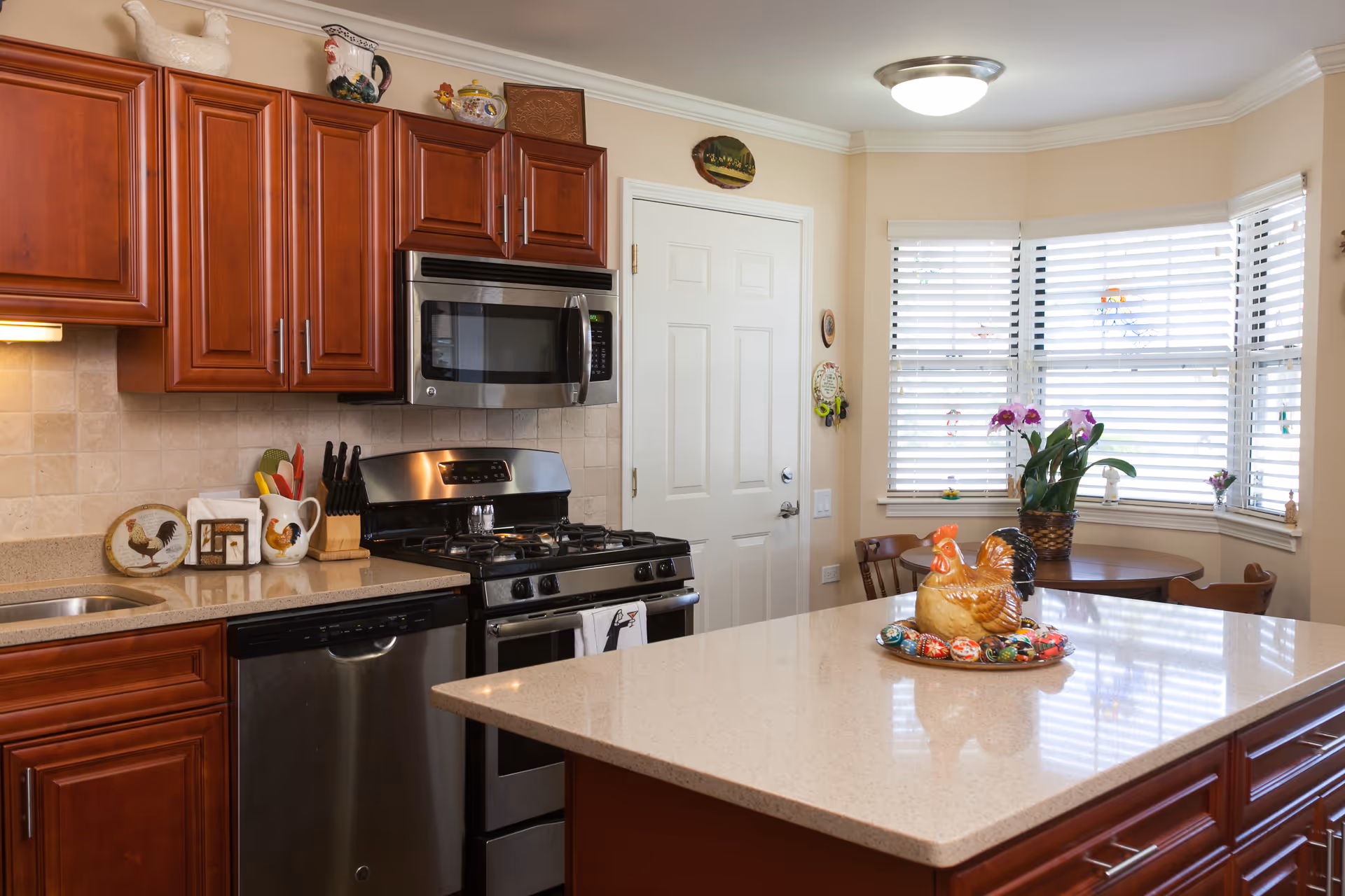 Bright kitchen with cherry wood cabinets, stainless steel appliances, a large island topped with a ceramic hen centerpiece, and a breakfast nook by a bay window.