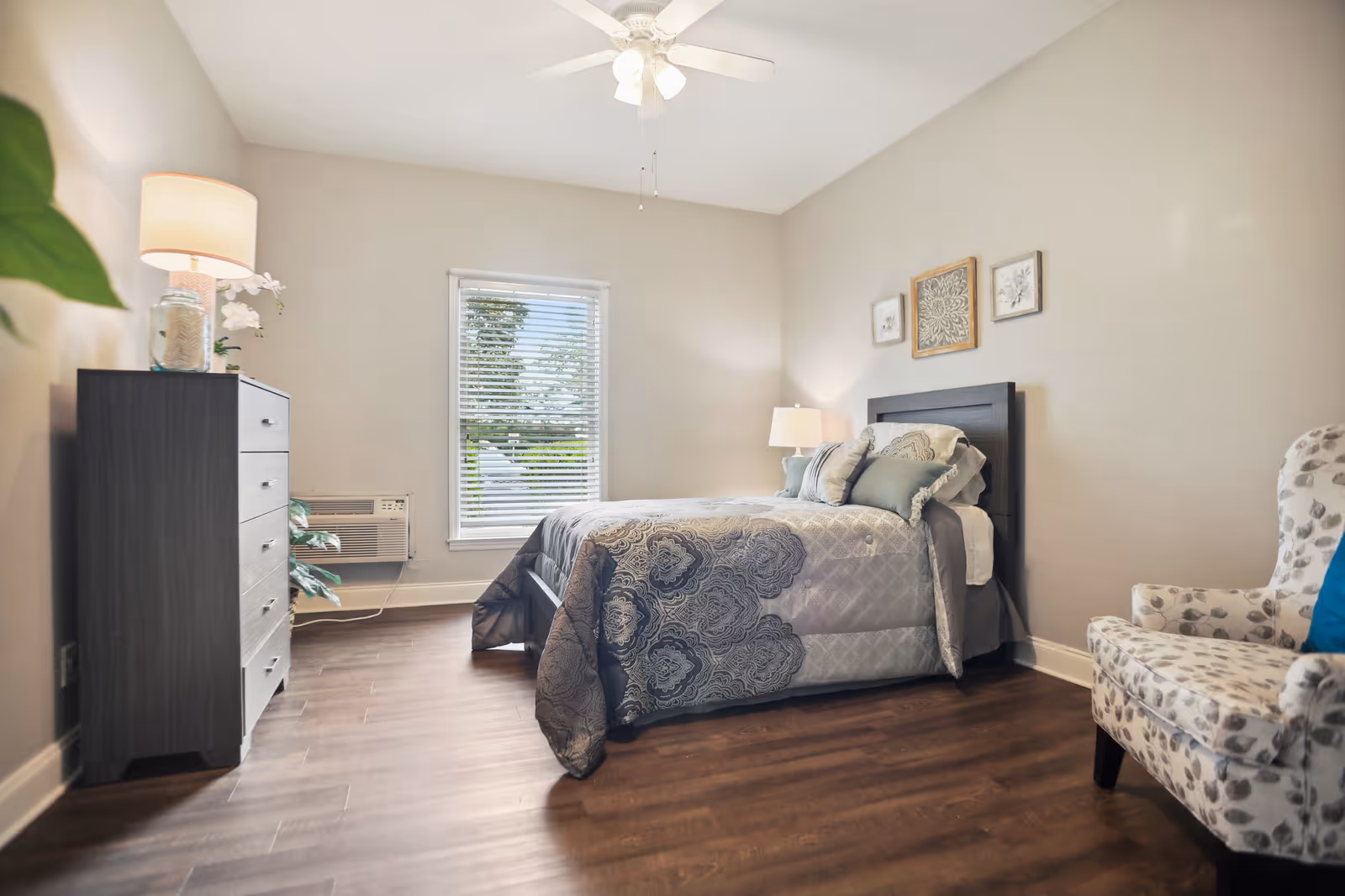 Furnished bedroom with a bed, bedside lamp, dresser, patterned armchair, window, and ceiling fan.