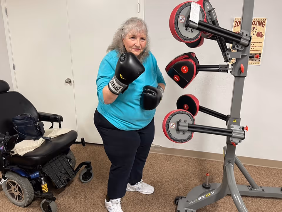 An elderly woman wearing black Everlast boxing gloves and a turquoise shirt is standing in a room next to a boxing training apparatus. There is a wheelchair with a bag on it to her left, and a boxing-themed poster on the wall behind her.