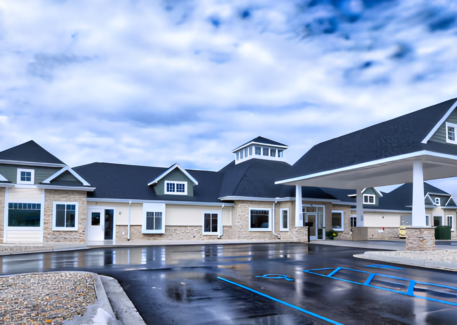 Front exterior of a single-story senior care building with a covered entrance/porte-cochere and a wet parking lot under a cloudy sky.
