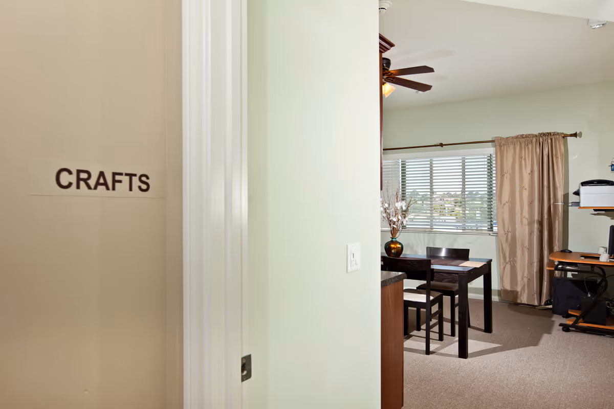 View into a crafts room with a table and chairs, a window with blinds and curtains, a ceiling fan, and a small desk area with a printer and office supplies.