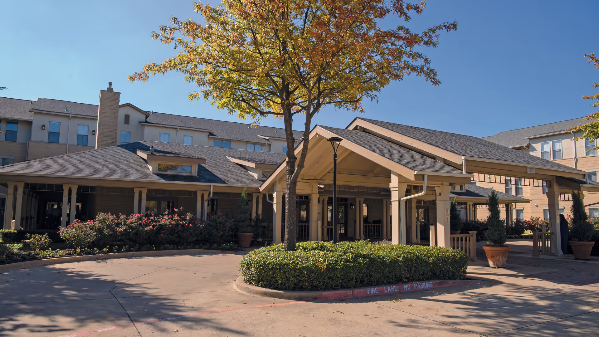 Exterior view of Town Village Crossing senior living facility showing the main entrance with a covered driveway, surrounded by landscaped bushes and a tree with autumn-colored leaves under a clear blue sky.