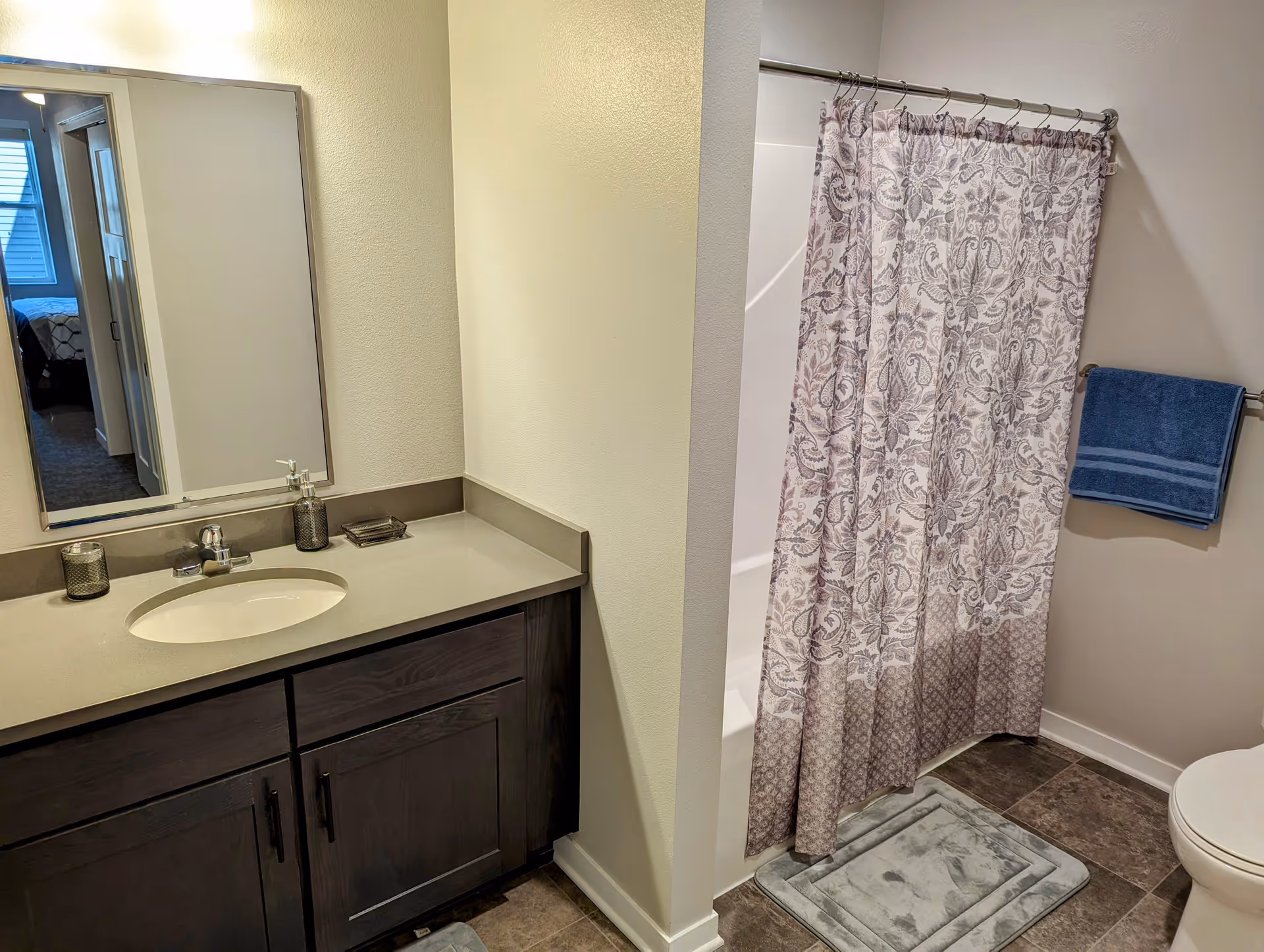 Bathroom with a dark wood vanity featuring a sink and countertop with soap dispenser and candle. A large mirror is mounted above the vanity. To the right, there is a shower with a patterned curtain, a blue towel hanging on a towel rack, and a toilet partially visible. The floor is tiled and there is a gray bath mat in front of the shower.