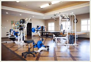 A spacious rehabilitation gym room with various exercise equipment including a stationary bike, weight machines, and a ping pong table. The room has large windows allowing natural light, a patterned wooden floor, and neutral-colored walls with ceiling lights.