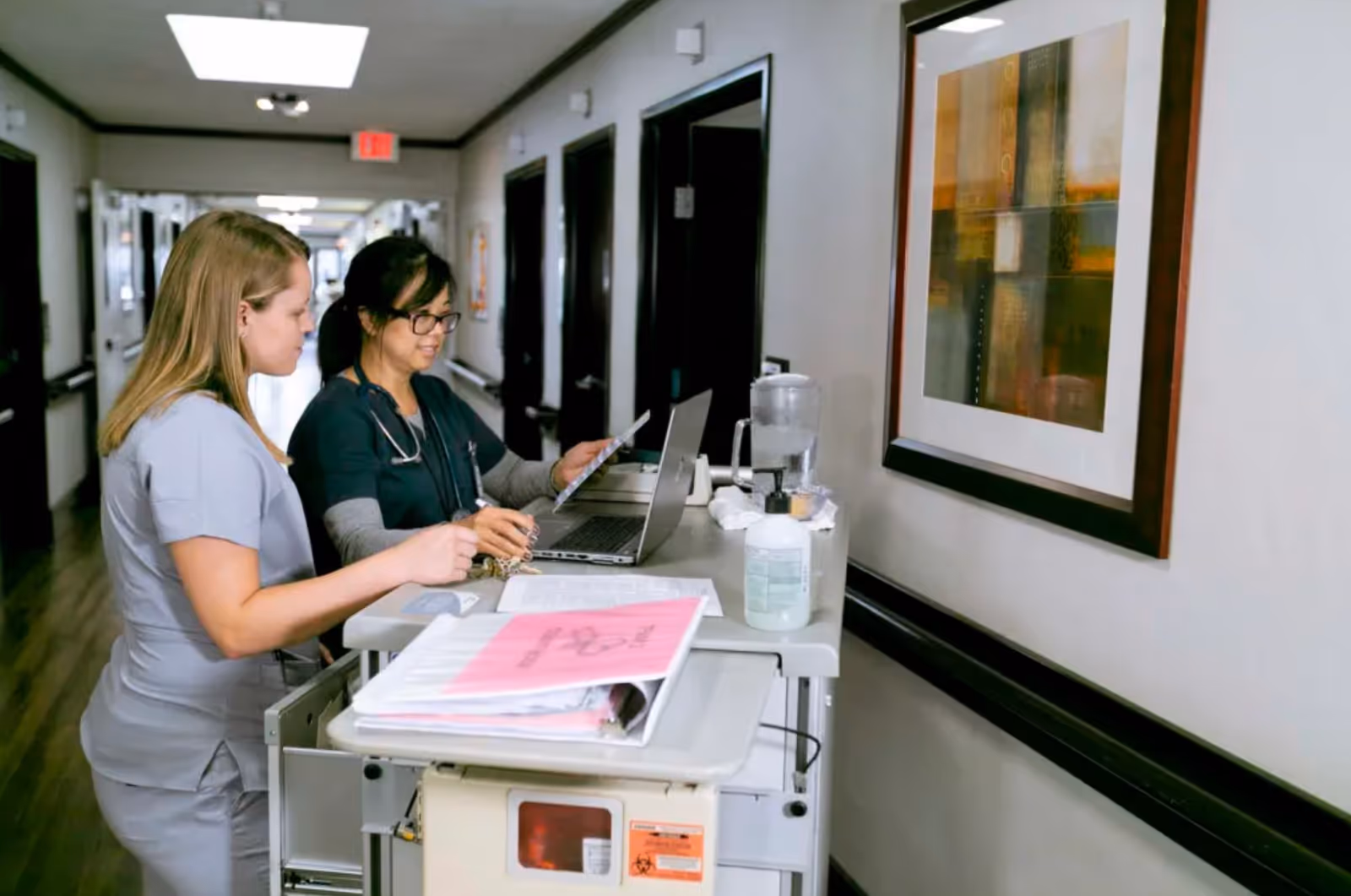 Two healthcare professionals standing in a hallway of a medical facility, reviewing documents and a laptop on a mobile workstation cart. The hallway has several doors and a framed abstract painting on the wall.