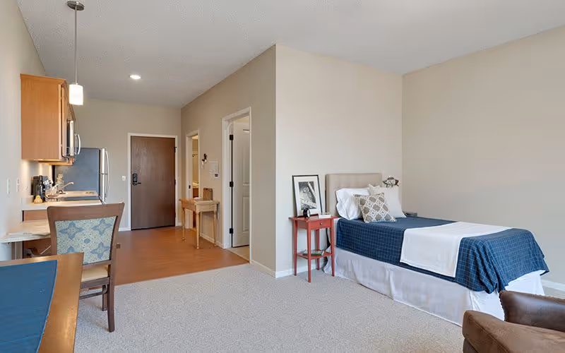 A senior living facility room featuring a single bed with blue and white bedding, a red bedside table with a framed picture and a lamp, a small kitchenette with wooden cabinets, a stainless steel refrigerator, and a dining table with chairs. The room has beige walls, carpeted flooring near the bed, and wood flooring near the kitchenette and entrance door.