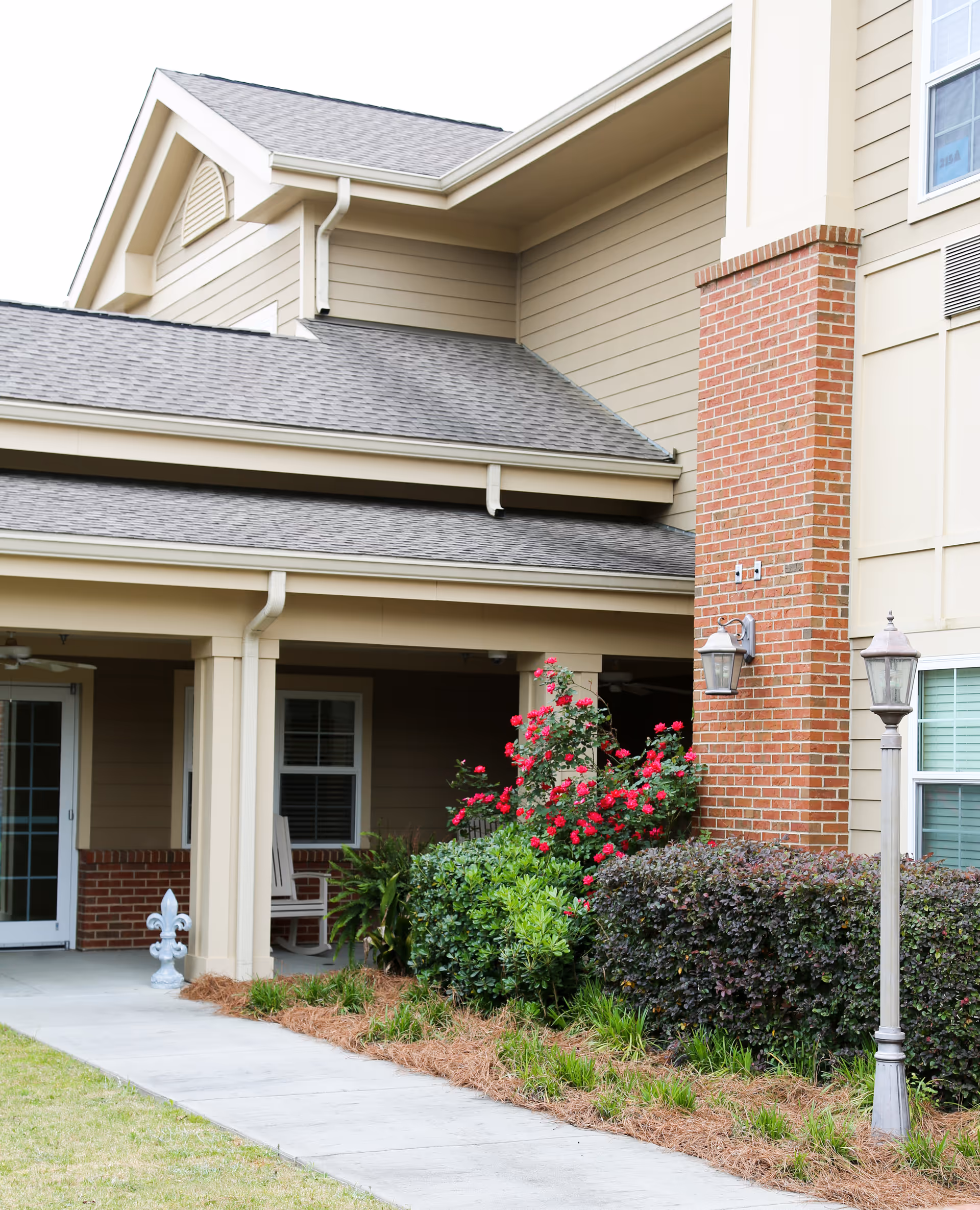 Covered entrance and porch of a beige building with a brick chimney, lamp posts, a walkway, and landscaped shrubs with red flowers.