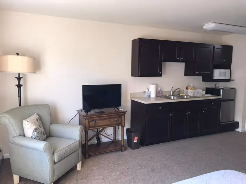 Studio living area with an armchair and TV beside a small kitchenette with dark cabinets, sink, microwave and mini-fridge.