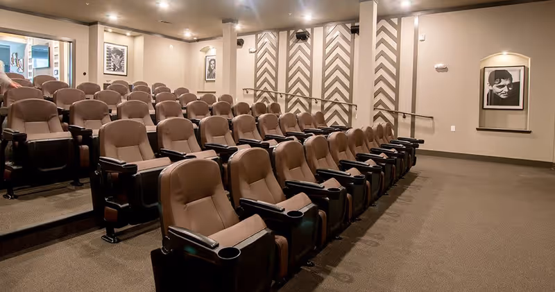 A small theater room with multiple rows of comfortable brown recliner chairs arranged in tiers facing a screen. The walls are decorated with black and white framed portraits and patterned panels. The room is well-lit with recessed ceiling lights.