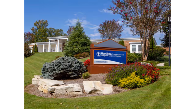 Exterior view of Ridgewood Manor Health and Rehabilitation building with a landscaped garden featuring bushes, flowers, and rocks in front. A blue sign with white text reads 'Hamilton Long Term Care Ridgewood Manor Health and Rehabilitation' with the address '1111 Burkhart Road'. Trees and a clear blue sky are visible in the background.