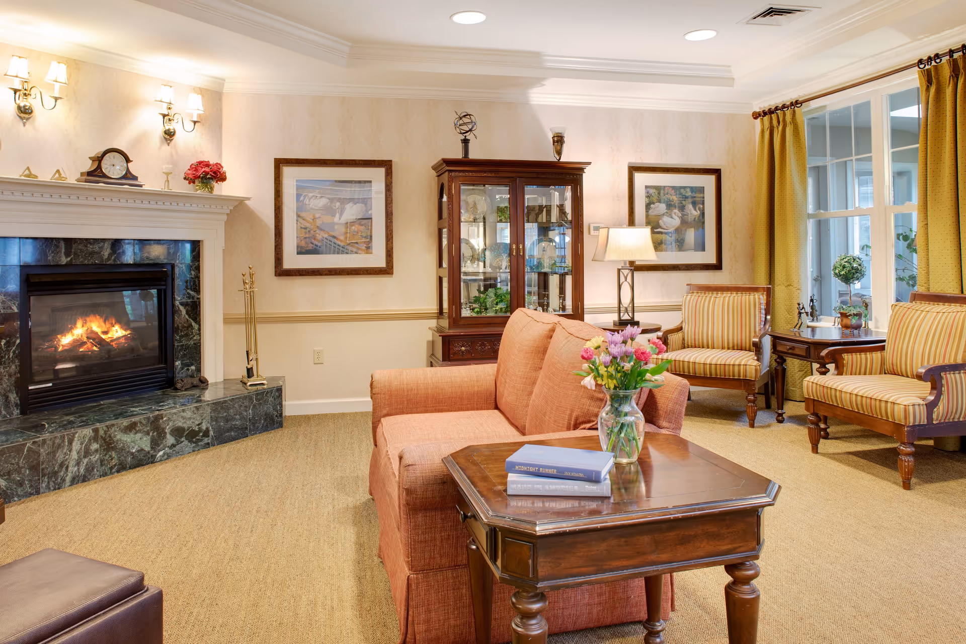 A cozy, well-lit living room with a lit fireplace, a sofa, a wooden coffee table holding a vase of flowers and books, striped armchairs, and a glass-fronted display cabinet.
