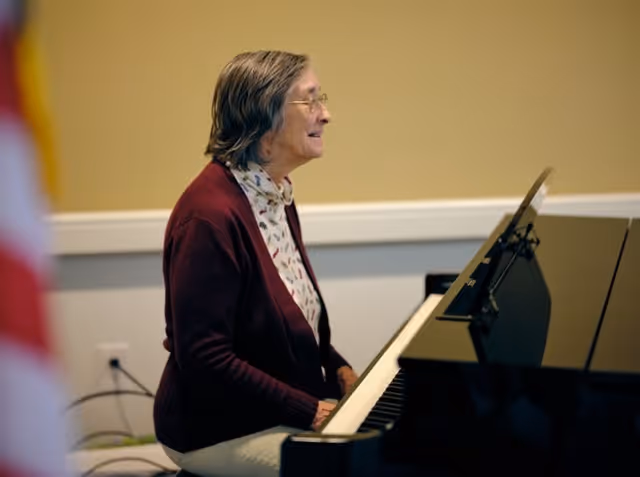 An elderly woman wearing glasses and a maroon cardigan is playing a black piano in a room with beige walls and white trim.