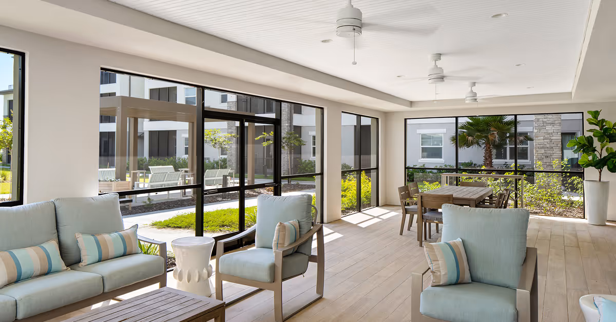 A bright and spacious indoor lounge area with light blue cushioned chairs and sofas, a wooden coffee table, and a dining table with chairs near large windows that offer a view of a landscaped outdoor courtyard with greenery and seating areas.