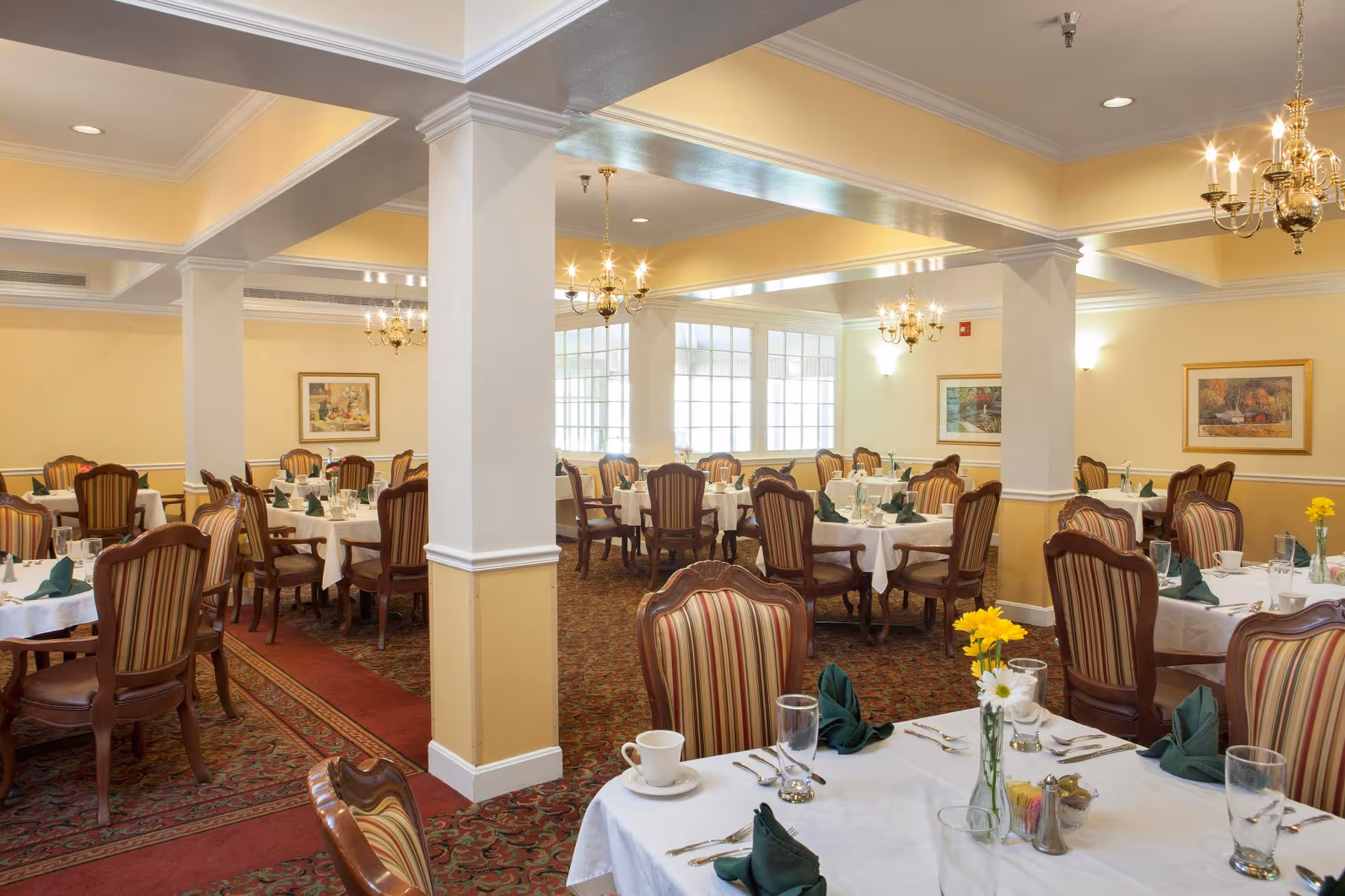 A spacious dining room with multiple round tables covered with white tablecloths, set with green folded napkins, glassware, and silverware. The room features striped upholstered chairs, chandeliers hanging from the ceiling, framed artwork on the walls, and large windows allowing natural light to fill the space.