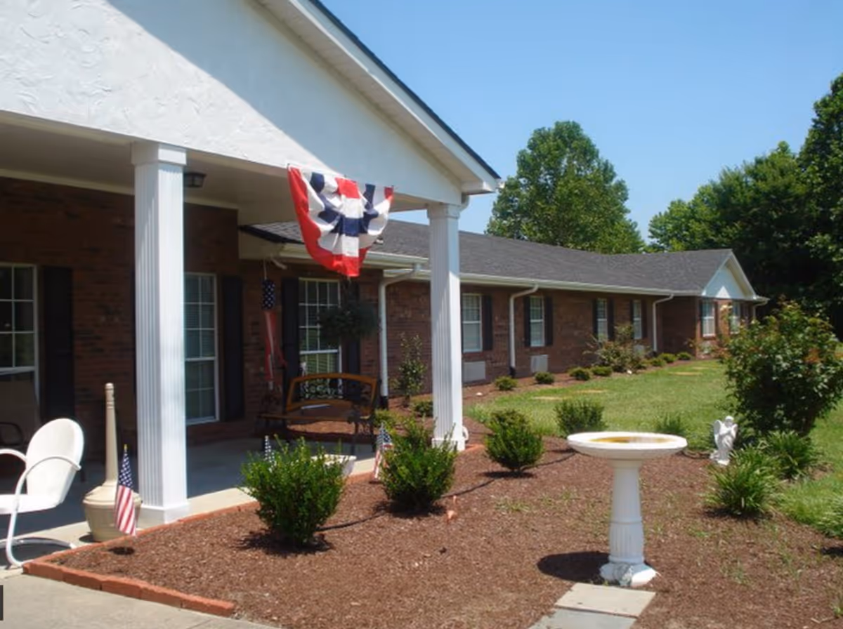 Front exterior of a single-story brick assisted living building with a covered porch, columns, American bunting and flags, a bench, and landscaped yard with a birdbath.
