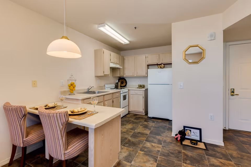 A small kitchen area with light wood cabinets, a white refrigerator, stove, and sink. There is a breakfast bar with two striped upholstered chairs, each set with a plate and wine glass. A pendant light hangs above the bar. The floor has dark tile, and there is a small pet feeding station with a framed sign and a toy near the entrance door.