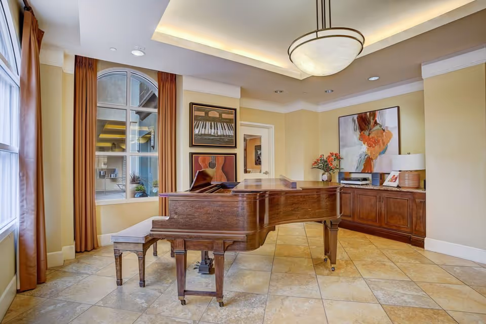 A bright room with a grand piano and matching bench on a tiled floor. The room has large windows with brown curtains, framed musical-themed artwork on the walls, a wooden sideboard with a lamp, flowers, and decorative items, and a modern ceiling light fixture.