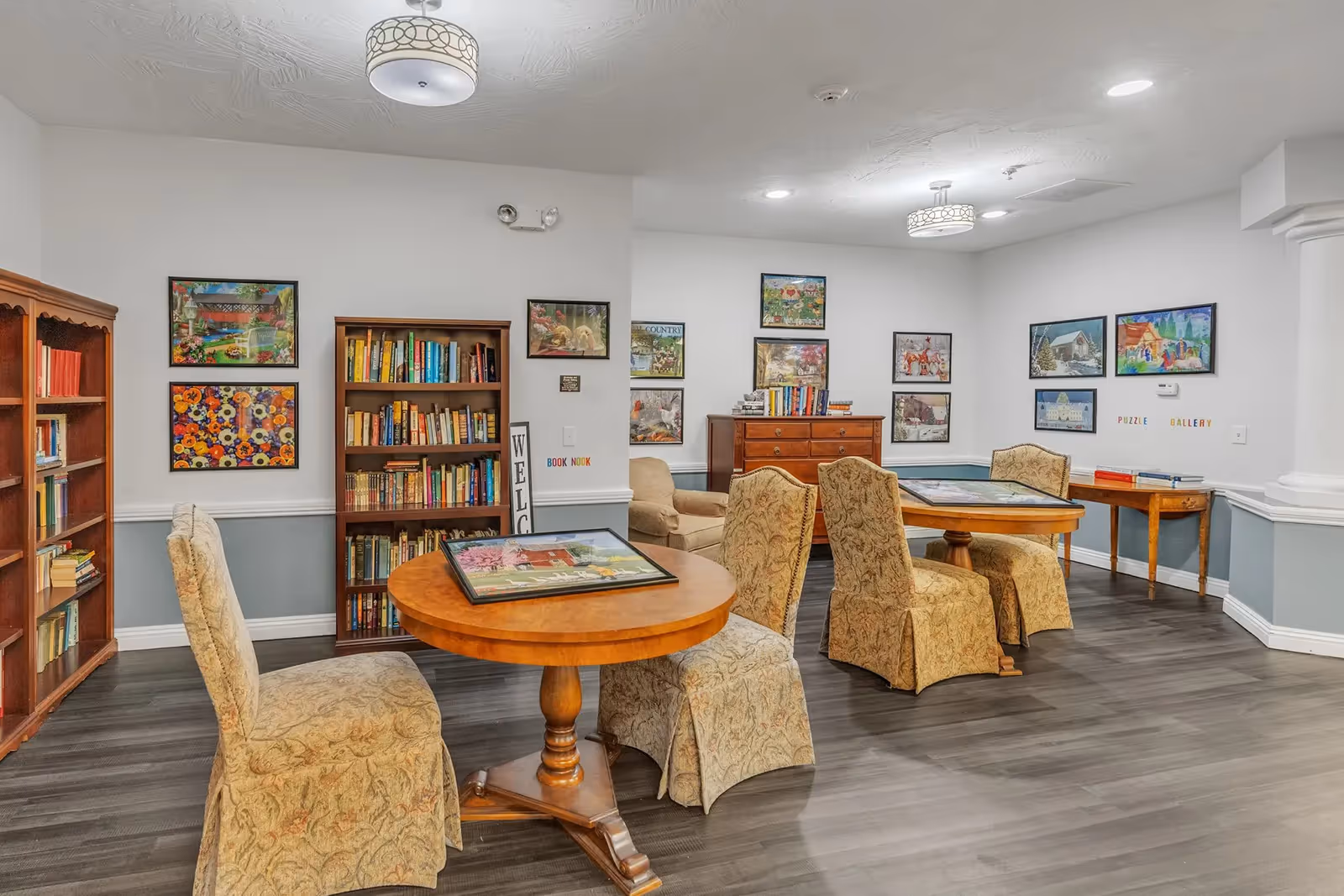 Well-lit common room with round wooden tables, upholstered chairs, bookshelves and framed artwork on the walls.