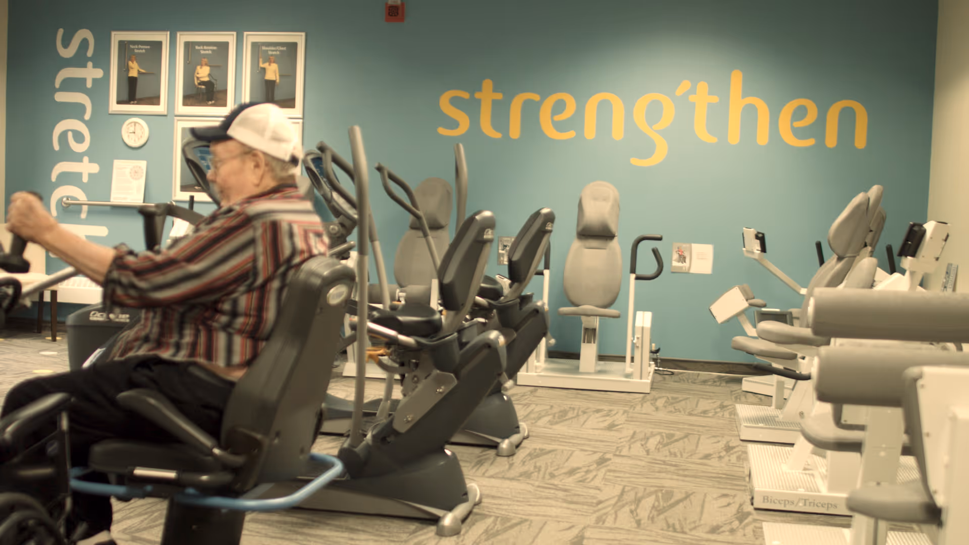 A person using a recumbent exercise machine in a fitness room filled with exercise equipment and a teal wall reading 'strengthen'.