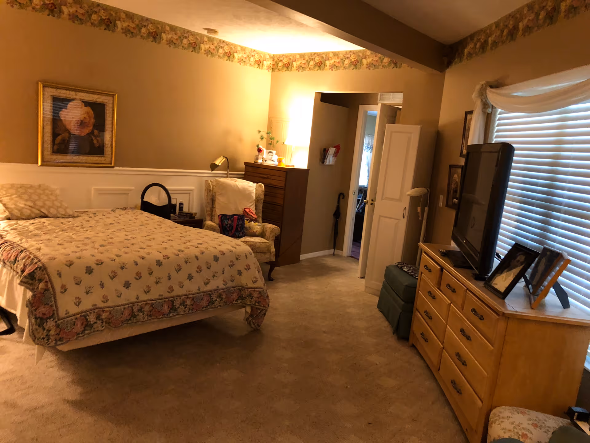 A spacious bedroom with a bed, armchair, dresser topped by a TV, and a window with blinds.