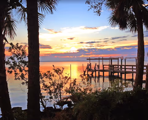 Sunset view over calm water with a wooden dock extending into the water, framed by palm trees and foliage in the foreground.