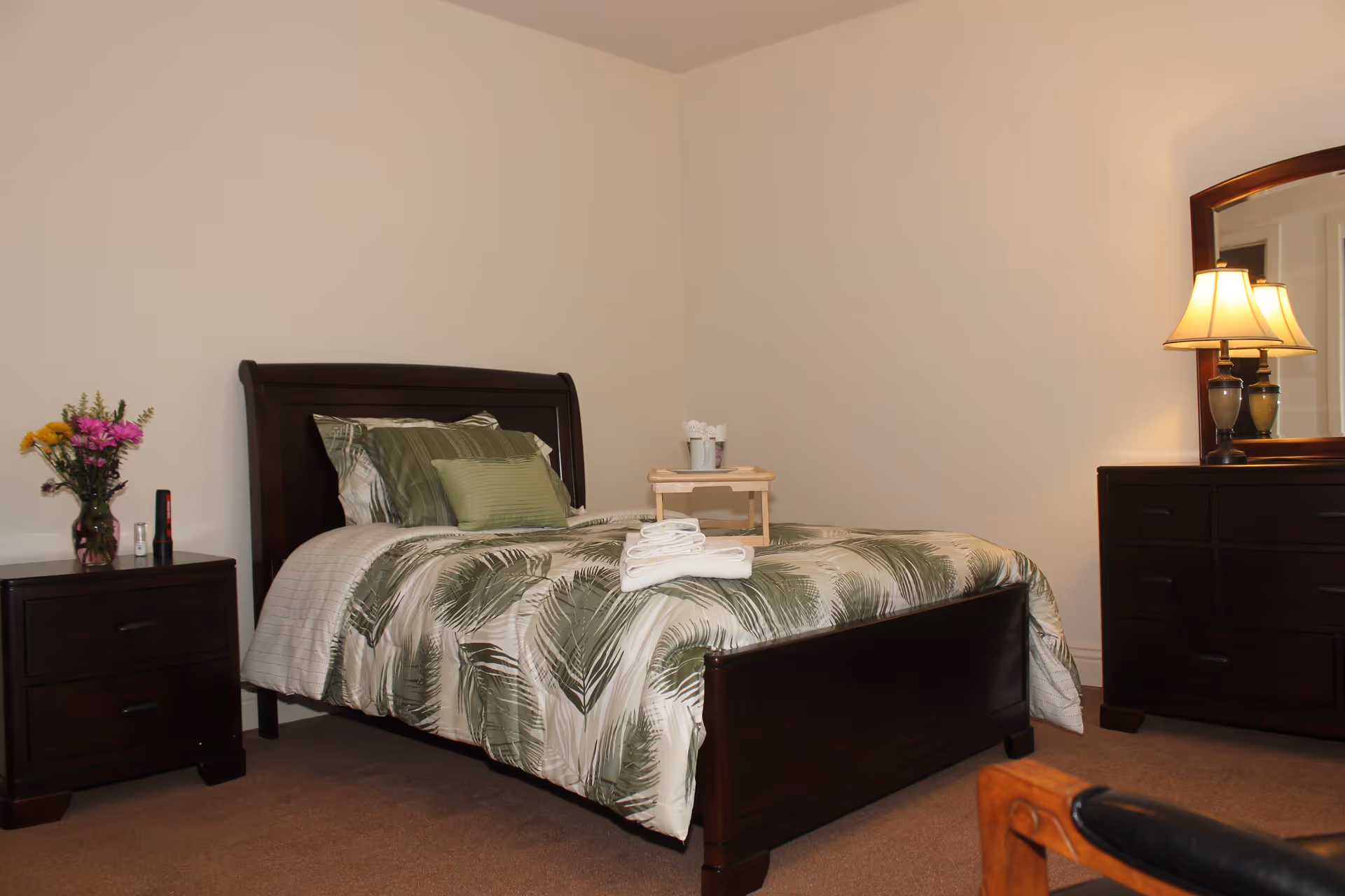 A bedroom with a dark wooden bed frame and a bed made with green and white leaf-patterned bedding. There are folded white towels on the bed and a small wooden tray with cups. To the left of the bed is a dark wooden nightstand with a vase of colorful flowers and a flashlight. To the right is a dark wooden dresser with a mirror and a lit table lamp. The walls are plain and light-colored, and the floor is carpeted.