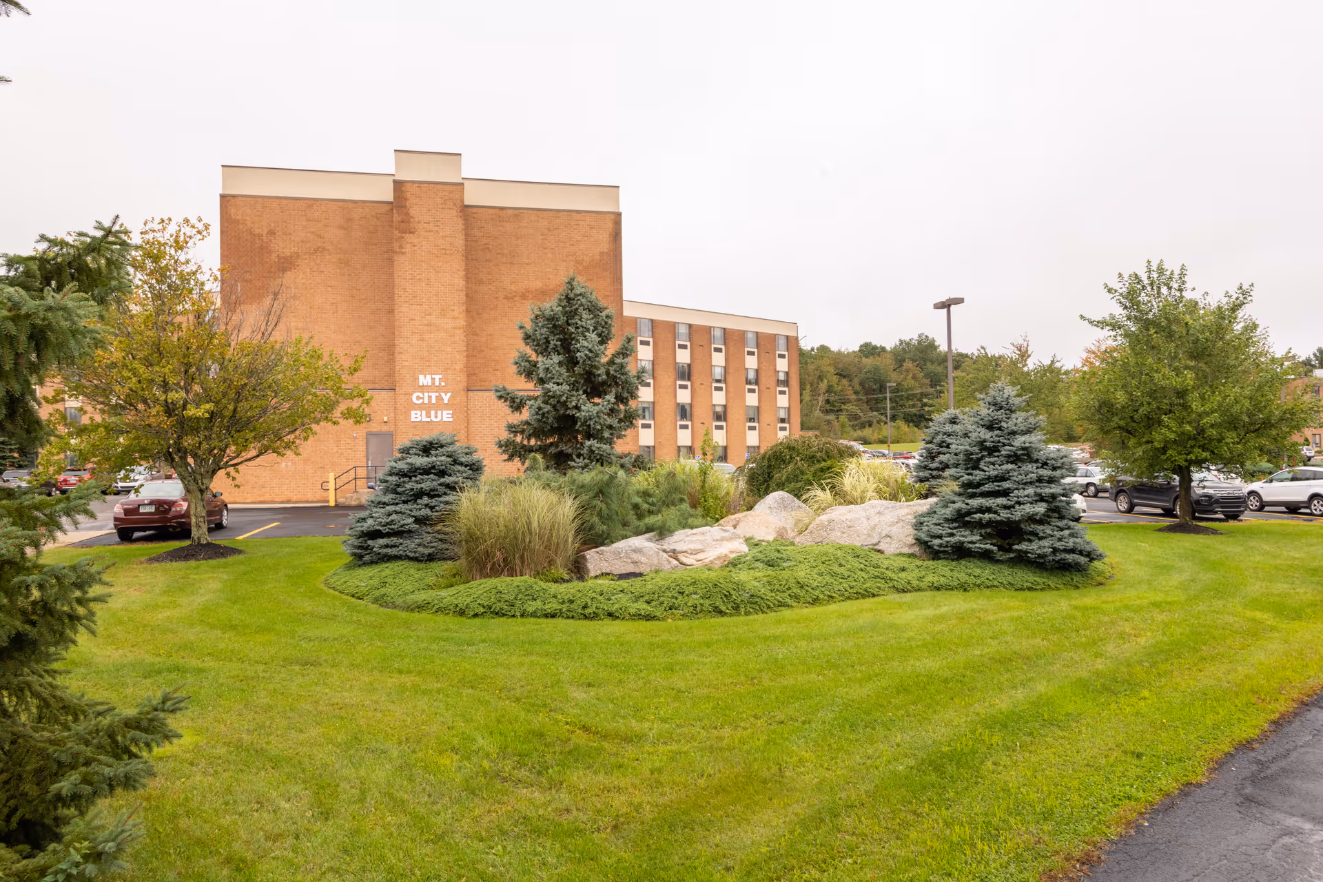 Brick nursing facility building with a landscaped lawn, rock garden, trees, and a parking lot.