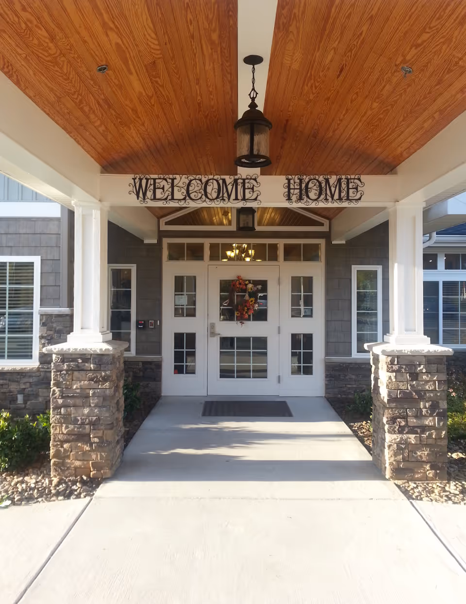 Entrance of a building with a covered porch featuring a wooden ceiling, stone pillars, and a white door decorated with a wreath. Above the door, a decorative sign reads 'WELCOME HOME'.