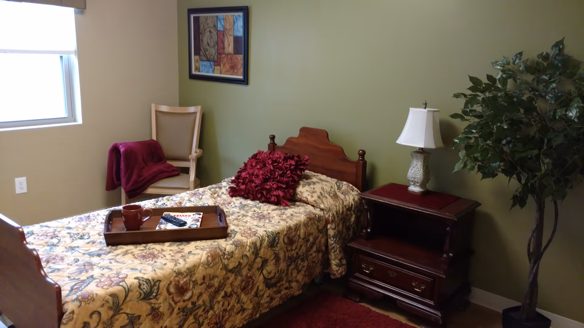A cozy bedroom with a single wooden bed covered in a floral bedspread and a red decorative pillow. A wooden tray with a cup, remote control, and magazine rests on the bed. Next to the bed is a wooden nightstand with a decorative lamp. A beige chair with a red blanket draped over it is positioned near a window. A framed abstract painting hangs on the green wall, and a tall leafy plant stands in the corner.
