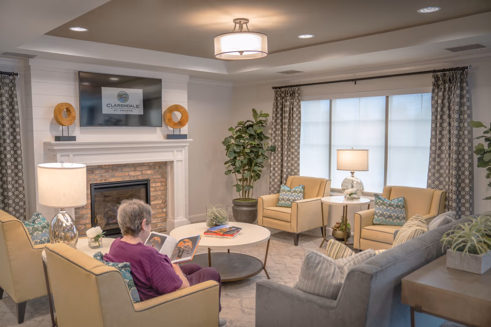 A cozy living room in Clarendale of St. Peters with a woman sitting on a yellow armchair reading a book. The room features a fireplace with a TV above it displaying the Clarendale of St. Peters logo, two yellow armchairs with patterned pillows, a gray sofa, a round coffee table, a side table with a lamp, large windows with patterned curtains, and potted plants.