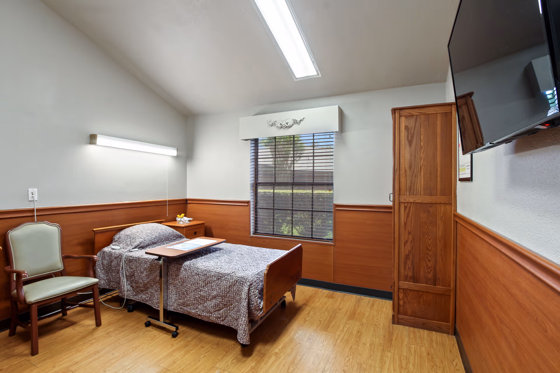 A small, clean patient room with a single hospital bed covered in a patterned blanket. Next to the bed is a wooden nightstand with a small tray table on wheels positioned over the bed. A green cushioned wooden chair is placed beside the bed. The room has wood paneling on the lower half of the walls and a large window with blinds letting in natural light. A flat-screen TV is mounted on the wall opposite the bed, and a wooden wardrobe stands next to the window. The floor is light wood, and the ceiling has a fluorescent light fixture.