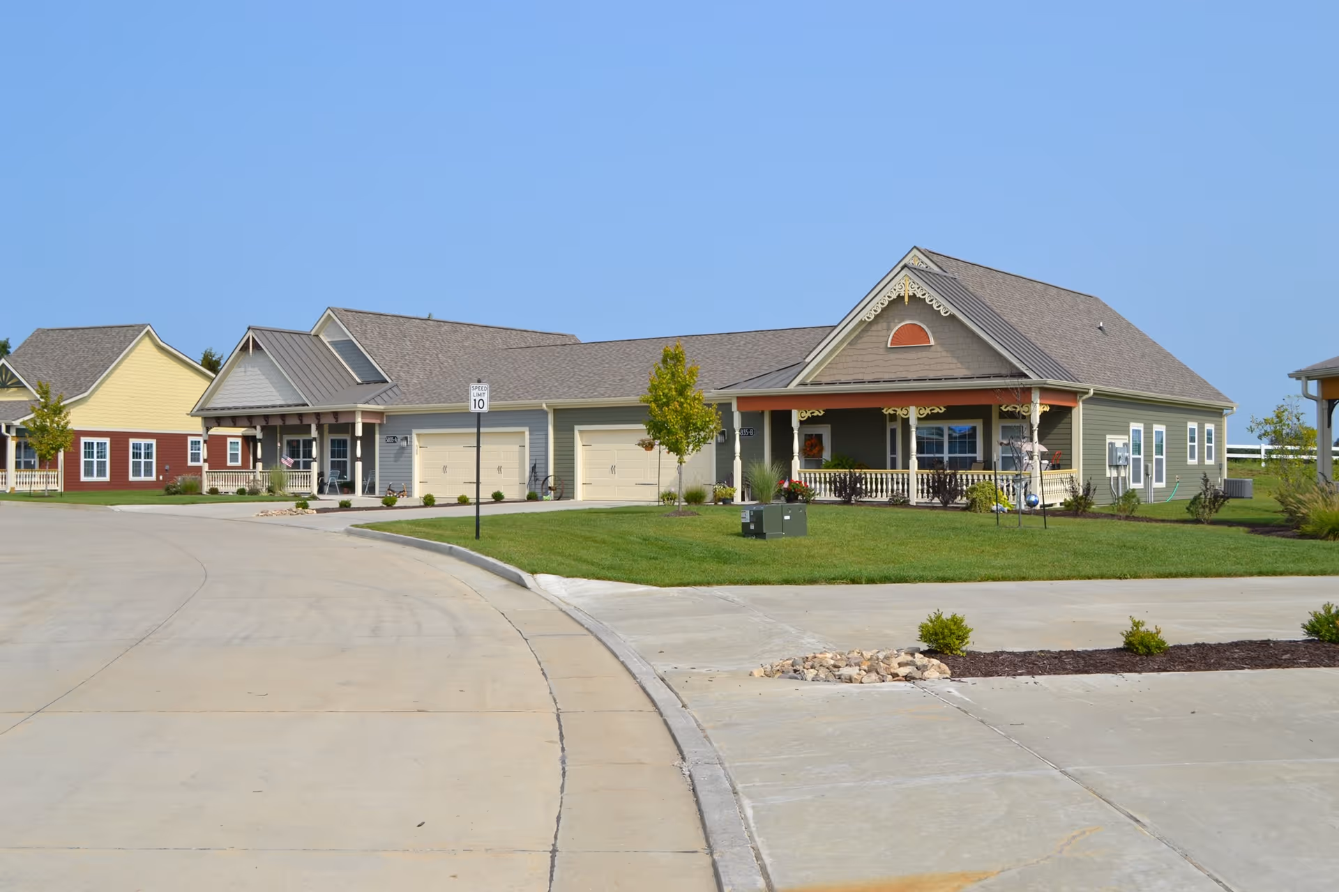 A row of single-story residential buildings with garages, porches, and well-maintained lawns under a clear blue sky, located along a curved concrete road with a speed limit sign of 10 mph.