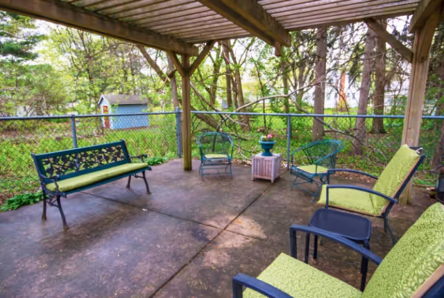 Covered outdoor patio with green-cushioned chairs and benches under a wooden pergola overlooking a fenced yard and trees.