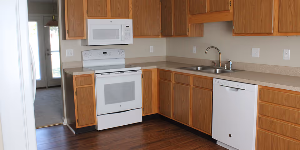 A kitchen with wooden cabinets, a white electric stove with an oven, a white microwave above the stove, a double sink with a faucet, and a white dishwasher. The floor is dark wood, and there is a door with glass panels leading to another room in the background.