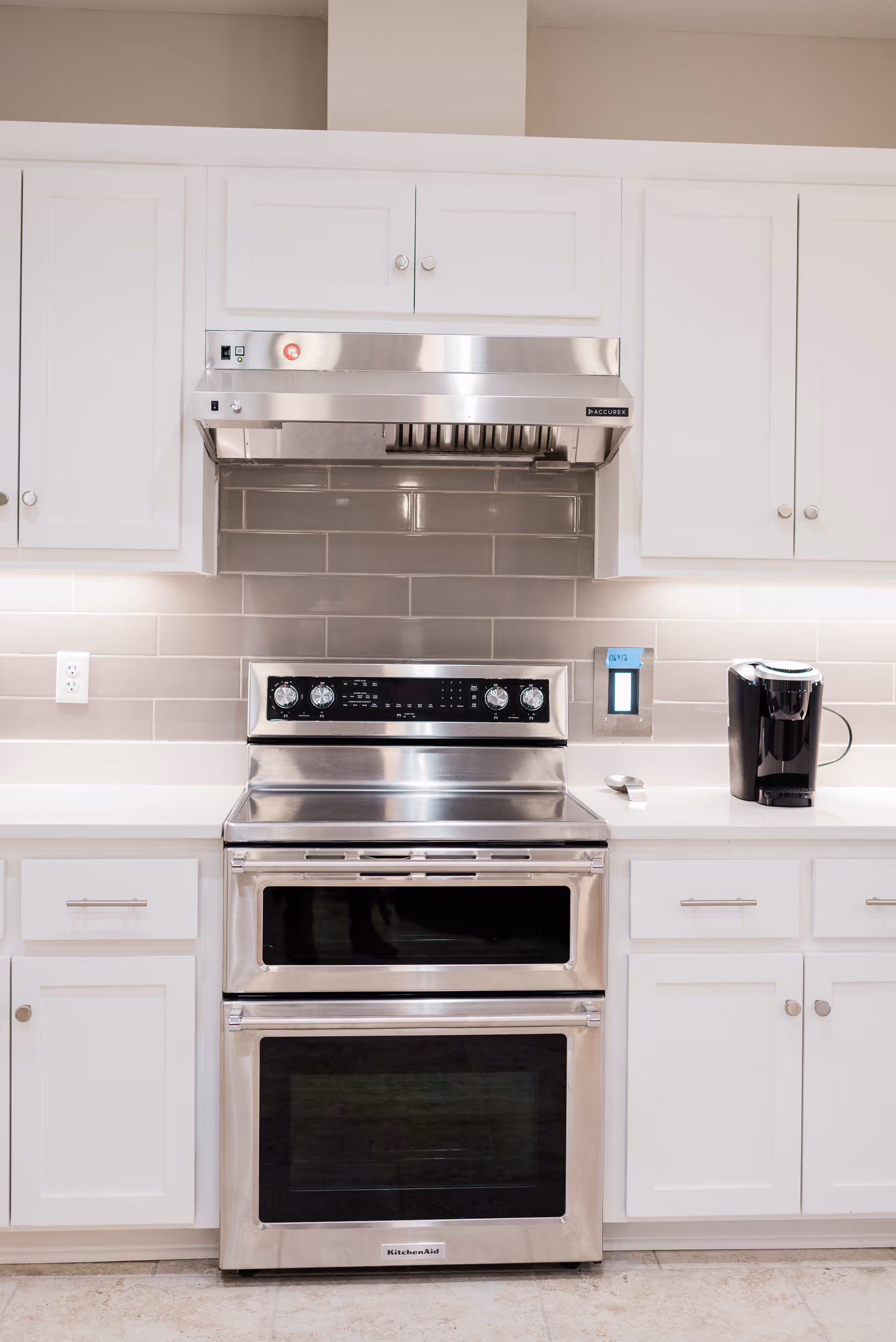 Modern kitchen with white cabinets, a stainless steel double oven stove, a stainless steel range hood, and a black coffee maker on the white countertop. The backsplash is made of glossy beige subway tiles.