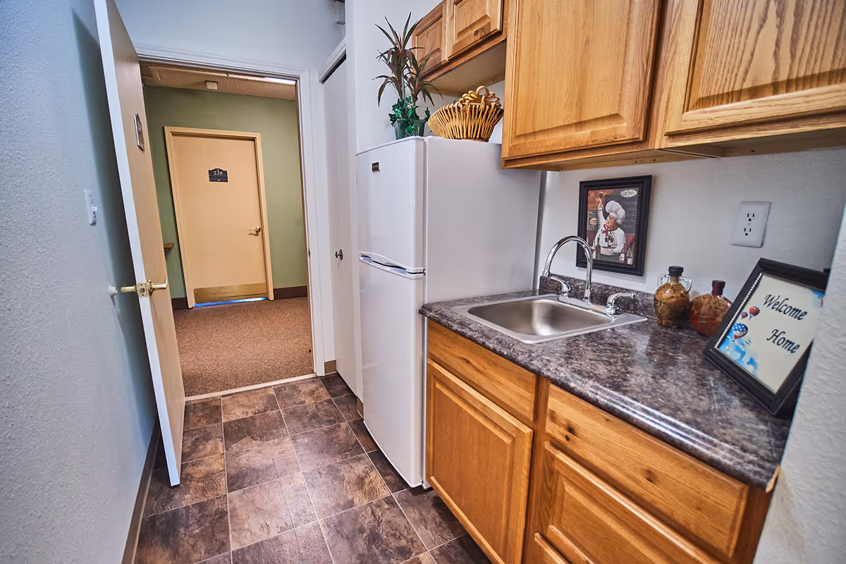 Small kitchenette with a sink, marble-look countertop, wooden cabinets and a refrigerator opening onto a hallway and door.