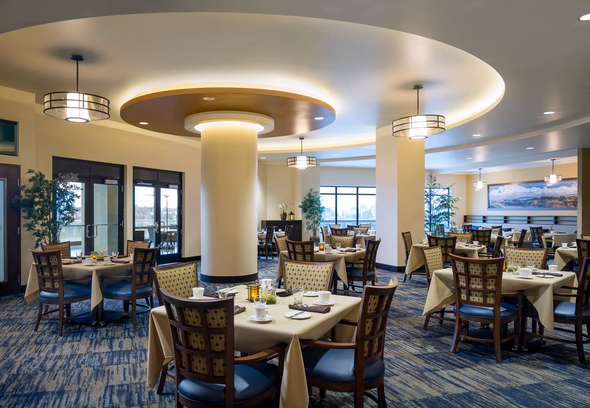 A spacious and well-lit dining room with multiple tables covered in beige tablecloths, each set with cups, plates, and napkins. The room features modern circular ceiling lights, large windows allowing natural light, and decorative plants near the walls.