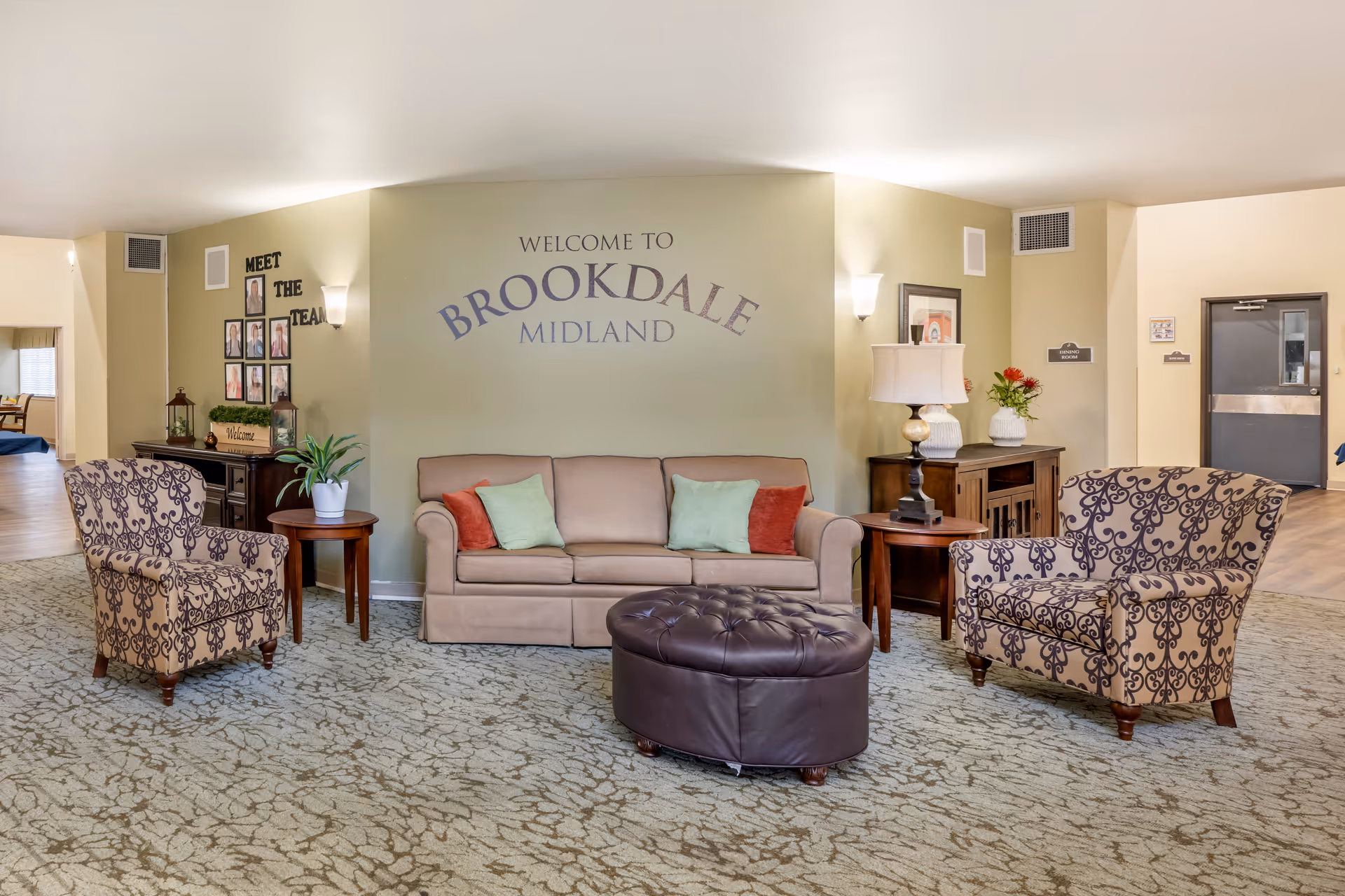 A welcoming seating area inside Brookdale Midland featuring a beige sofa with red and green pillows, two patterned armchairs, a round dark brown tufted ottoman, and side tables with plants and a lamp. The wall behind the sofa has the text 'Welcome to Brookdale Midland' and a 'Meet the Team' photo display on the left side. The area has carpeted flooring and soft lighting.