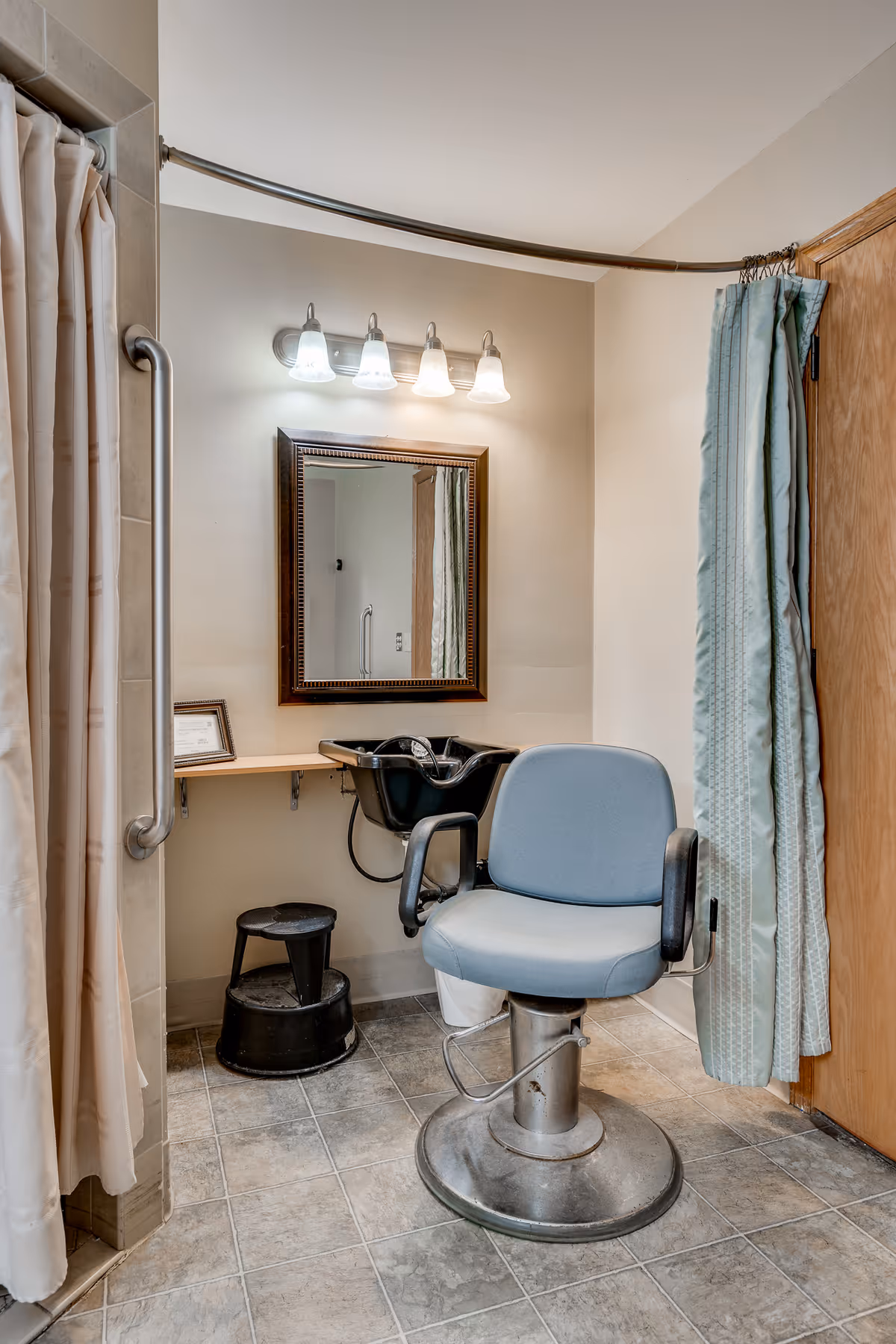 A hair-styling station with a padded swivel salon chair, sink basin, mirror, and overhead lights in a tiled interior room.
